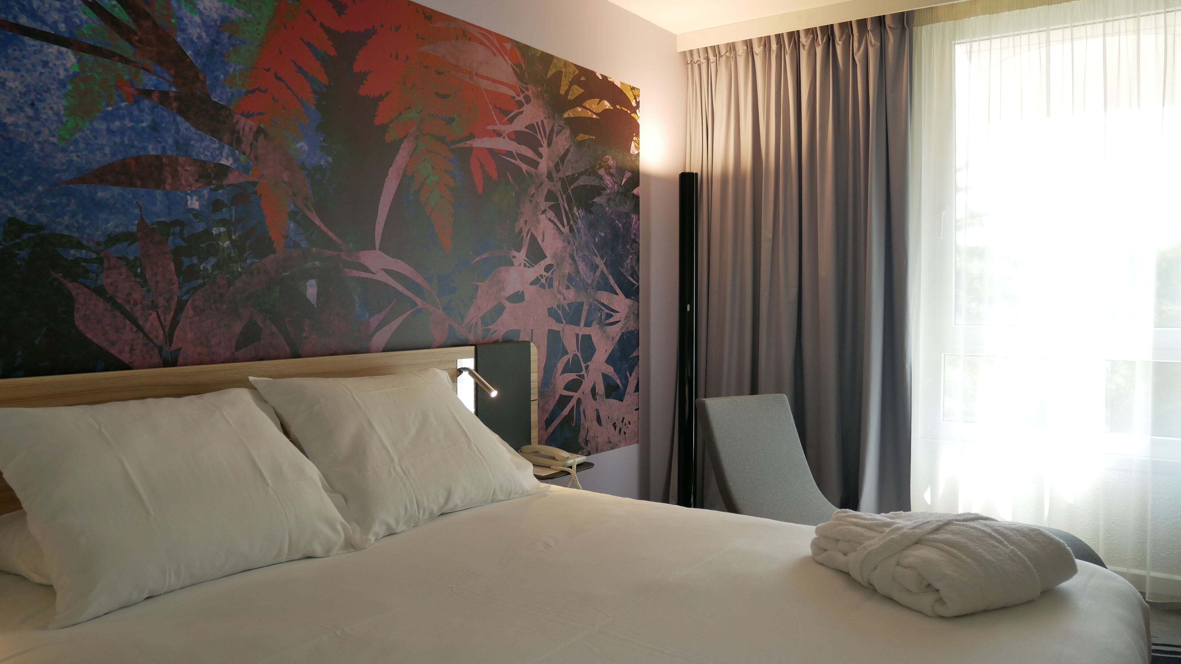A hotel room with a large bed, a chair, a painting on the wall, and a window with curtains.