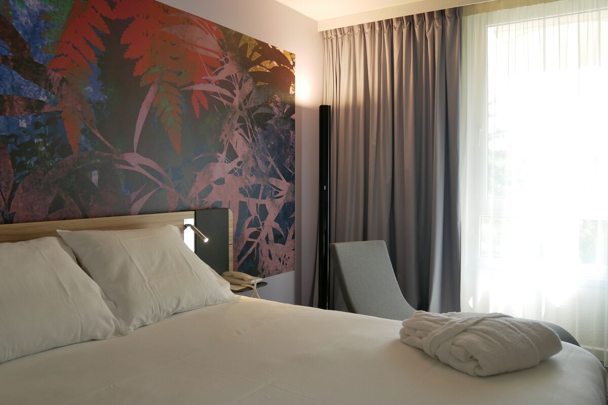 Habitación de hotel con una cama grande, una silla, un cuadro en la pared y una ventana con cortinas.