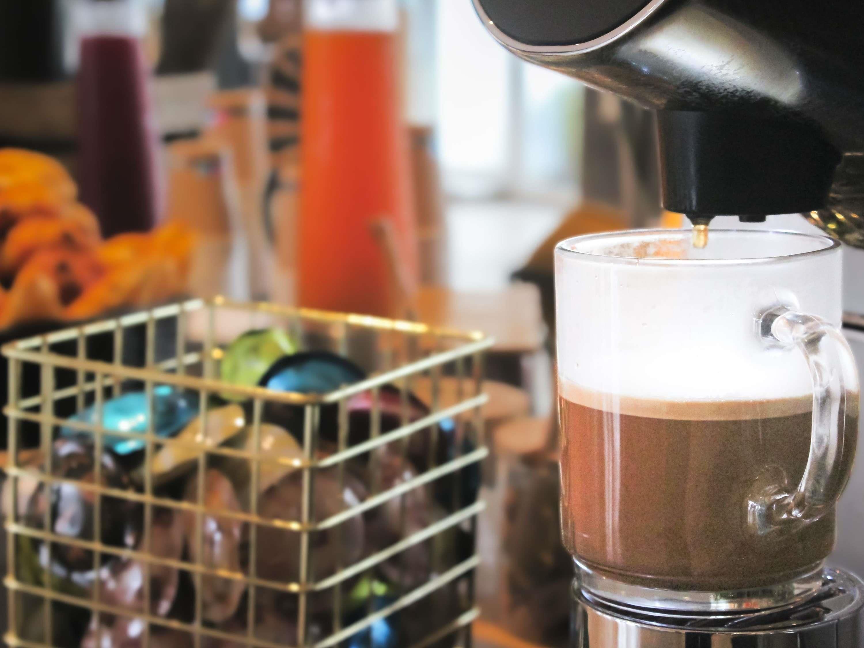 Una copa de vidrio transparente con una bebida en capas, una cesta metálica con botellas y una cafetera.