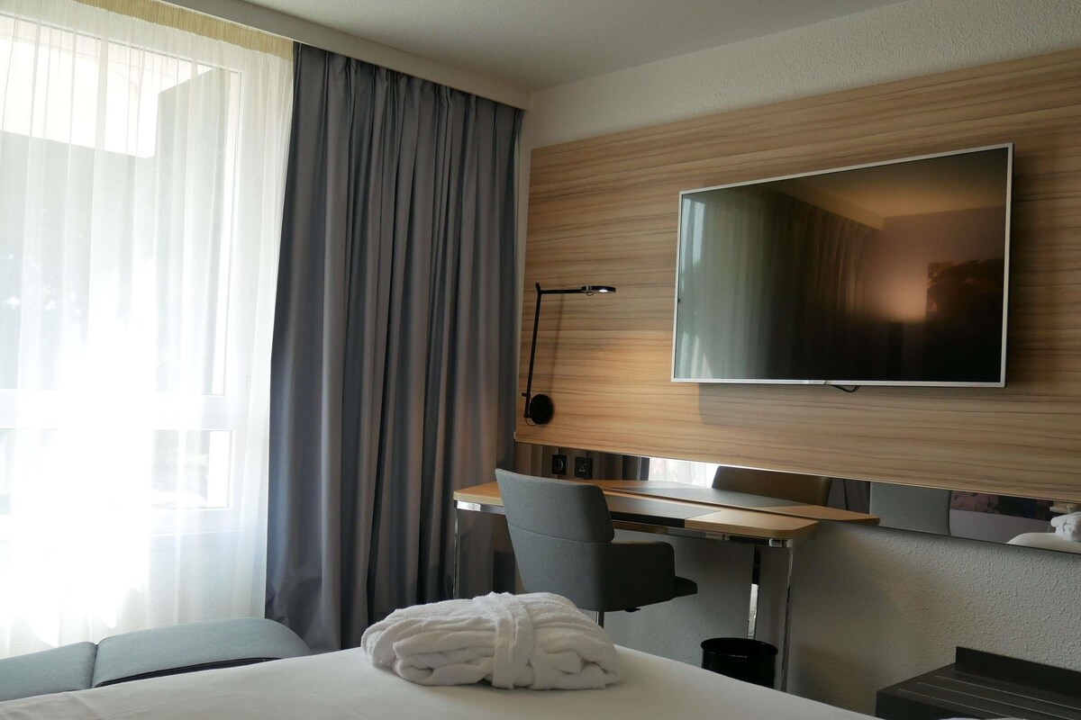 A hotel room with a bed, a desk, a chair, a TV, and a window with curtains.
