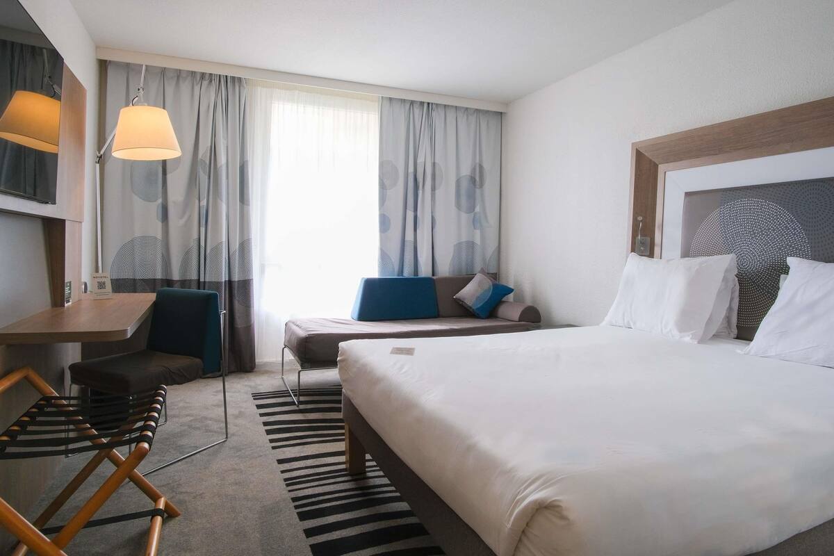 A hotel room with a bed, a desk, a chair, a sofa, and a window with curtains.