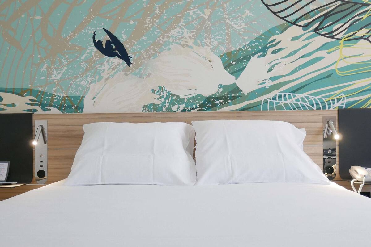 A hotel room with a large bed, white bedding, a wooden headboard, a wall mural, and a bedside table with a lamp and a phone.