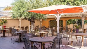 Un patio al aire libre con mesas y sillas redondas, rodeado de árboles y cubierto por un gran paraguas naranja.
