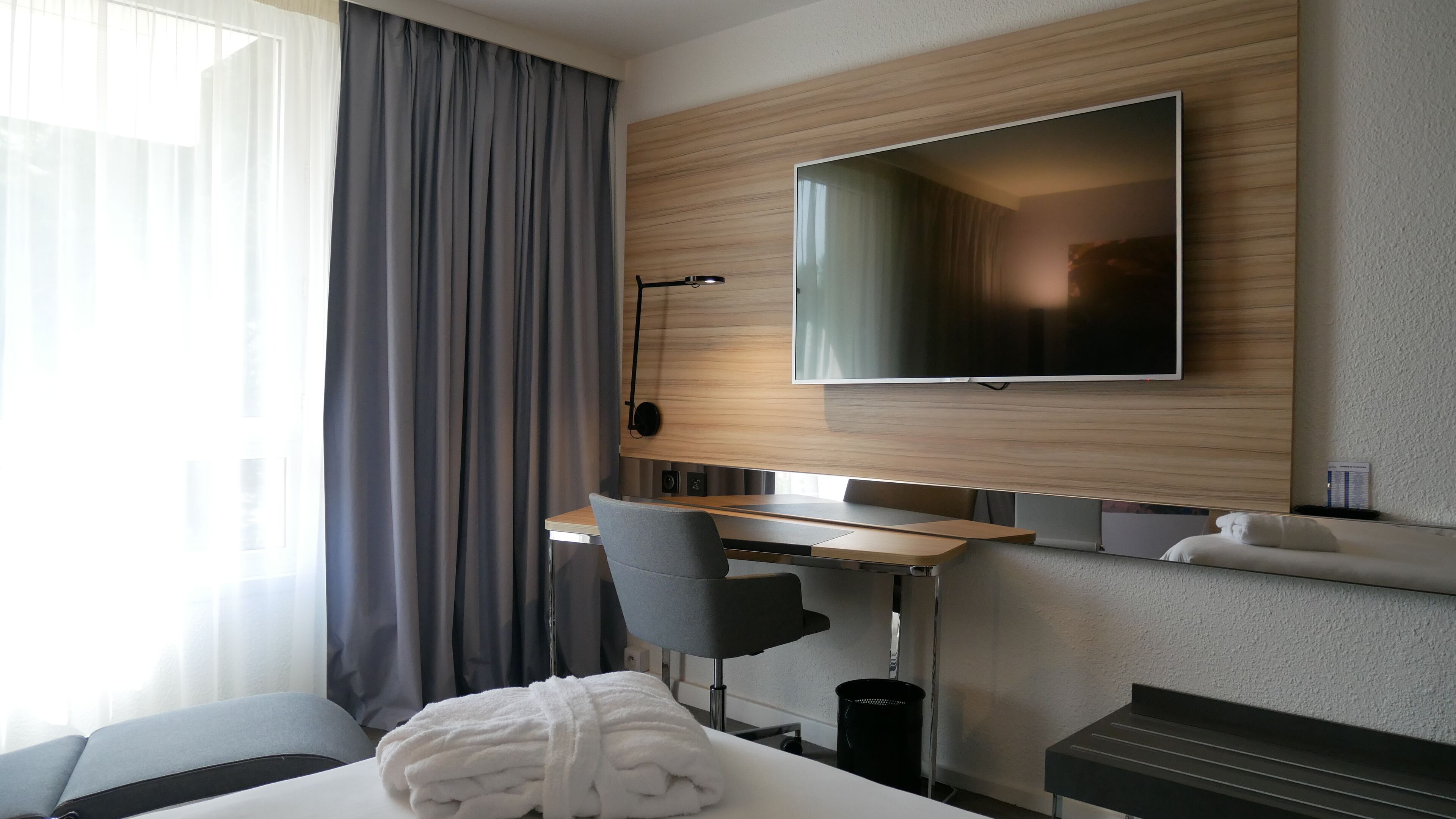 A hotel room with a bed, a desk with a chair, a TV mounted on the wall, and a window with curtains.