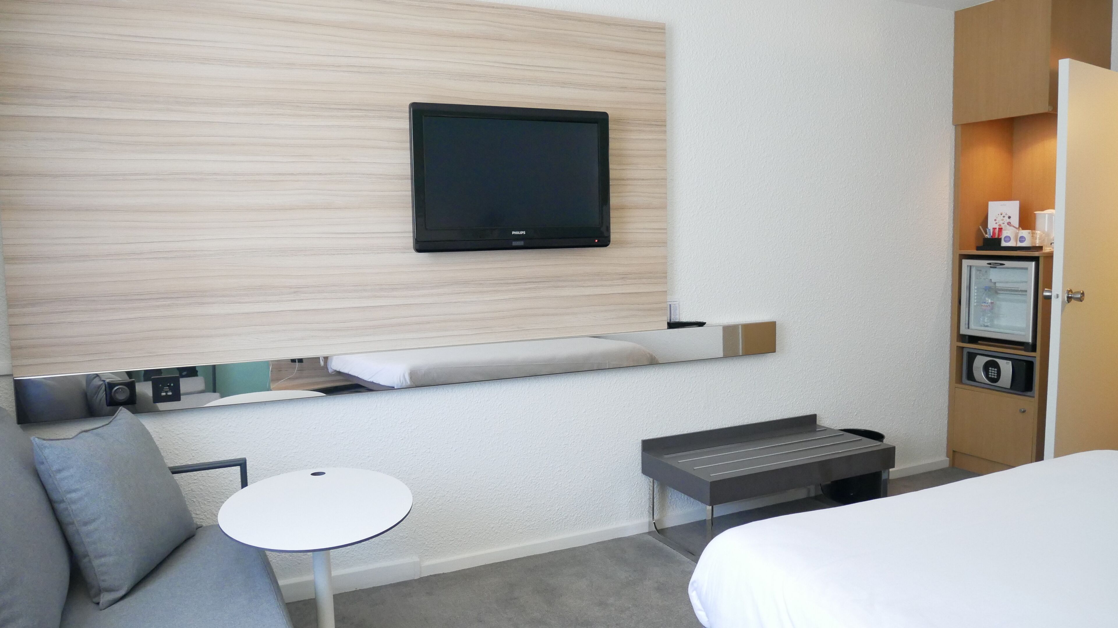 Habitación de hotel con cama, una pequeña mesa redonda, sofá, televisor montado en la pared y minibar.