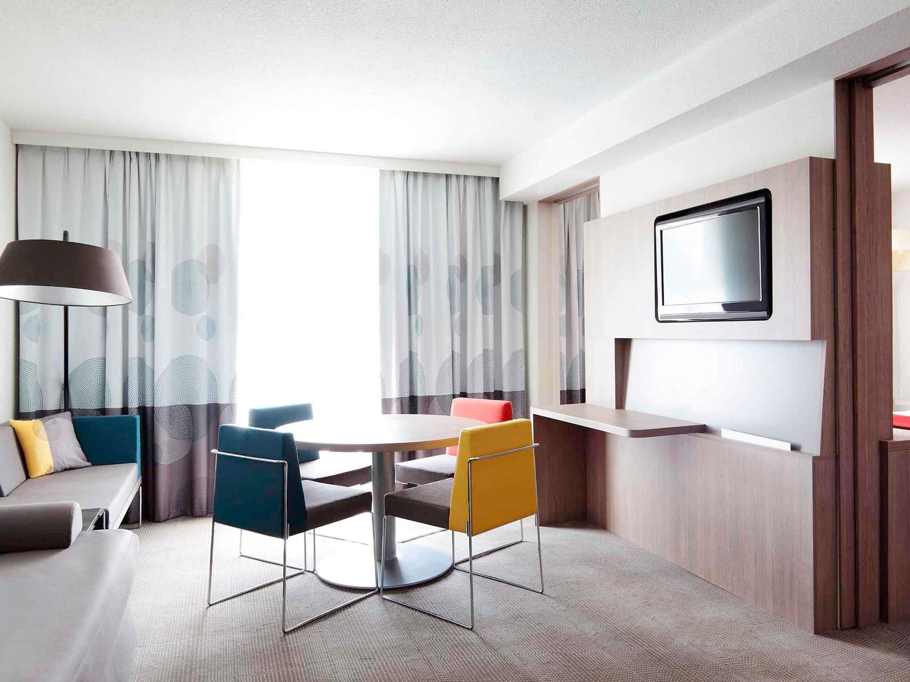 Una camera d'albergo moderna con un tavolo rotondo, due sedie, un divano, una TV a muro e un'ampia finestra con tende.