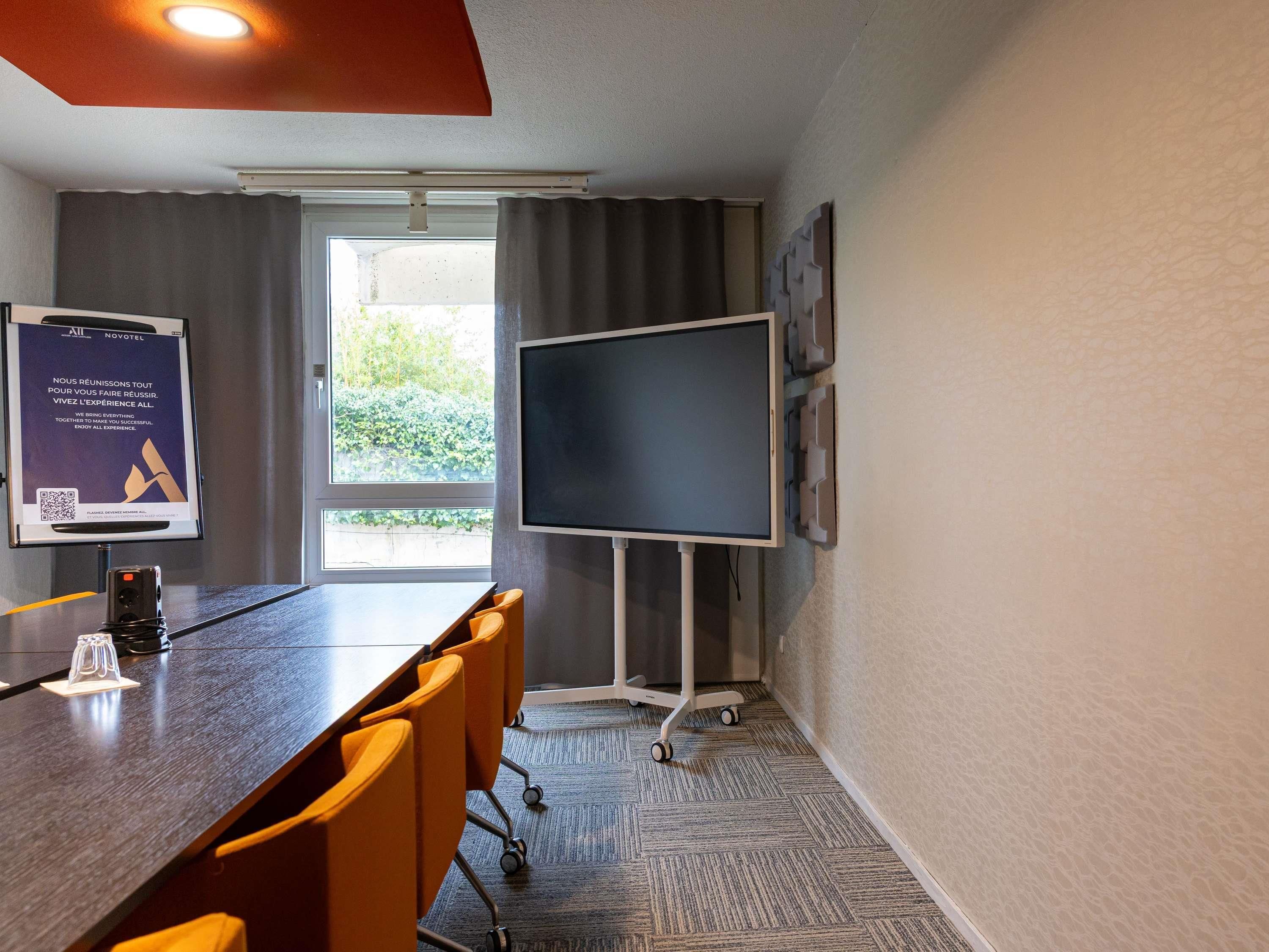 A conference room with a long table, chairs, a large screen, and a window with curtains.