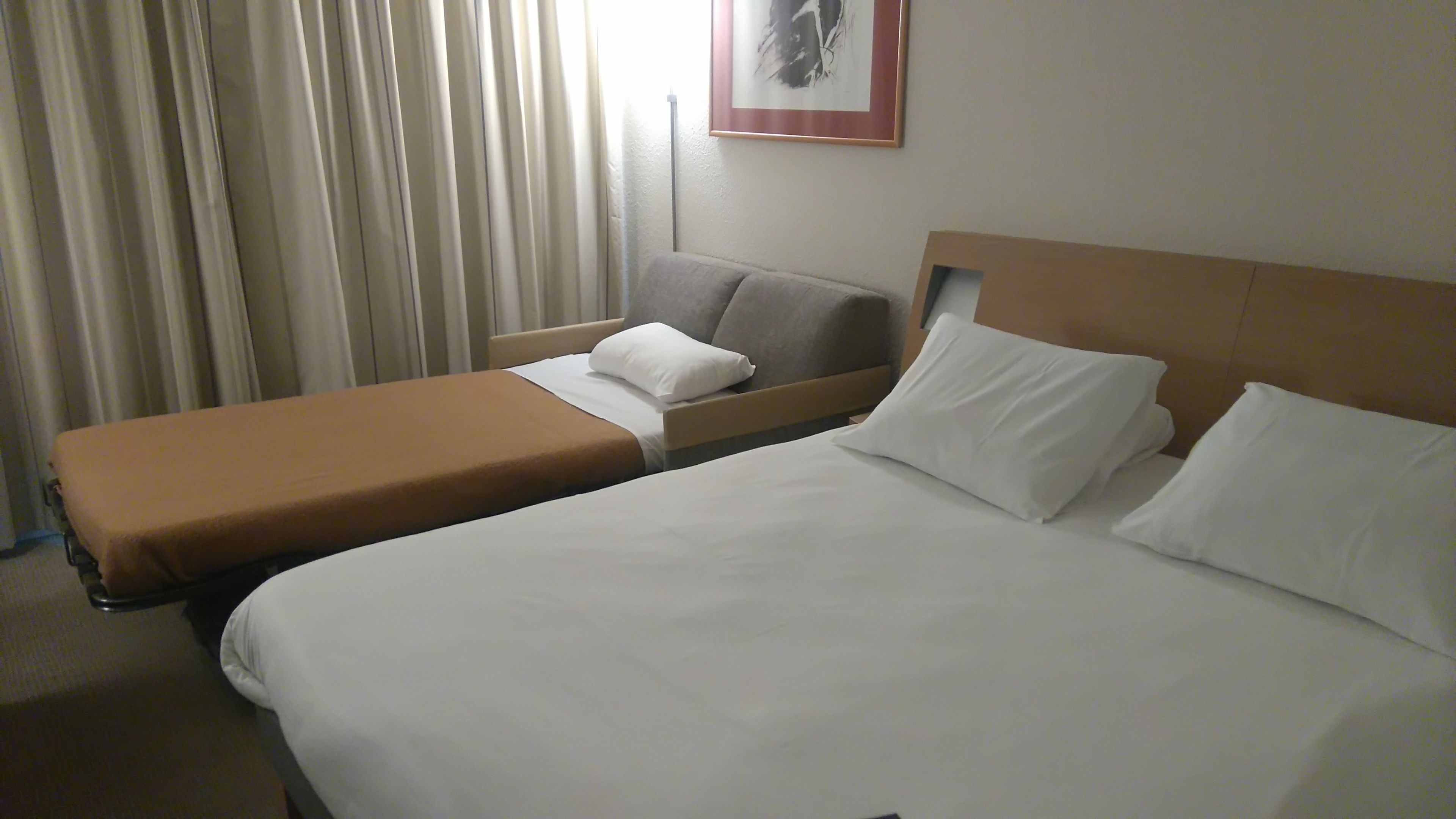 A hotel room with two beds, a sofa, and a large window with curtains.
