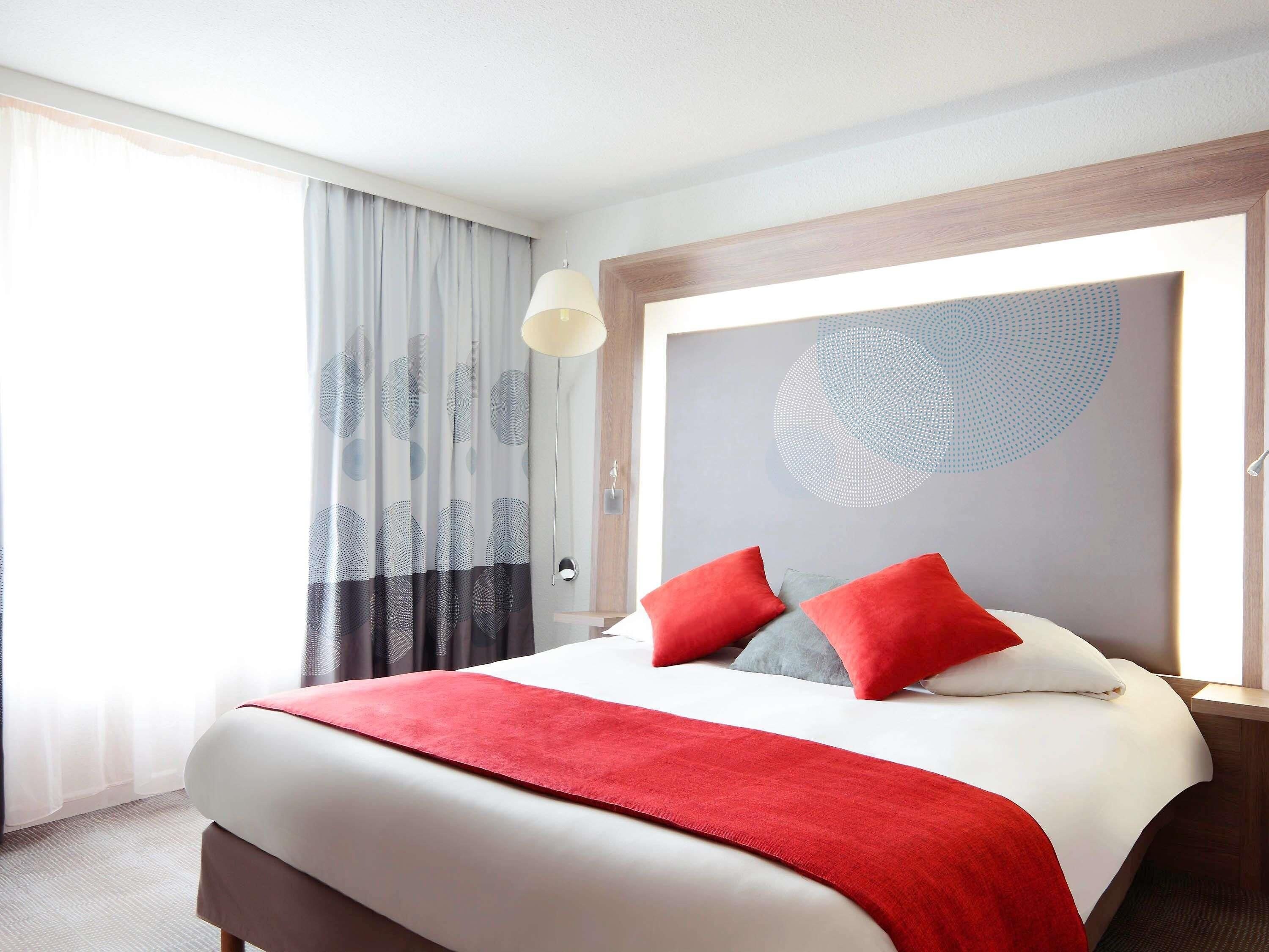 A hotel room with a large bed, a headboard with a circular design, a window with curtains, and a bedside table.