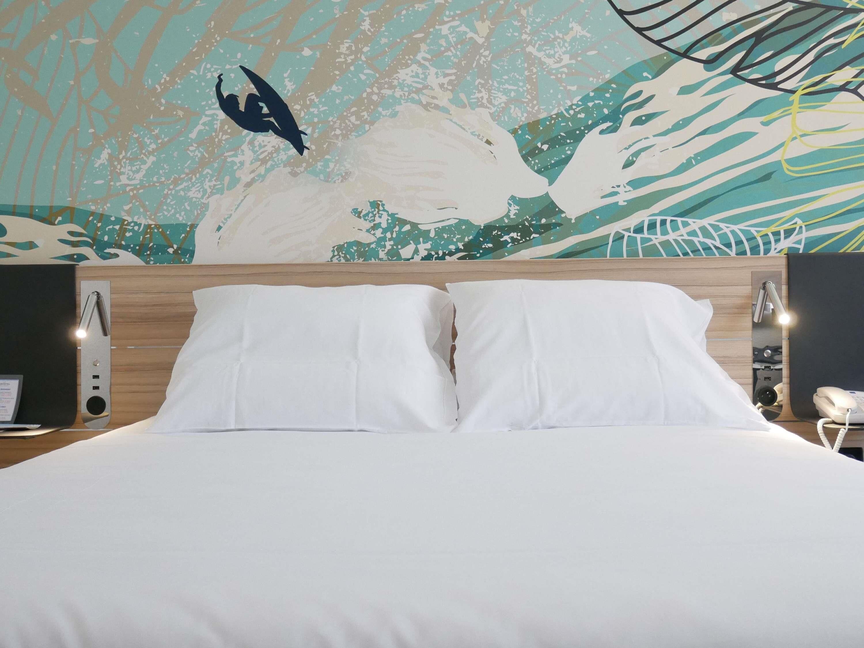 Una habitación de hotel con cama, almohadas blancas, cabecera de madera, un mural en la pared, una mesita de noche con lámpara y teléfono.
