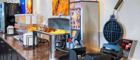Zona de buffet con una máquina para hacer gofres, una cafetera y diversas opciones de comida.