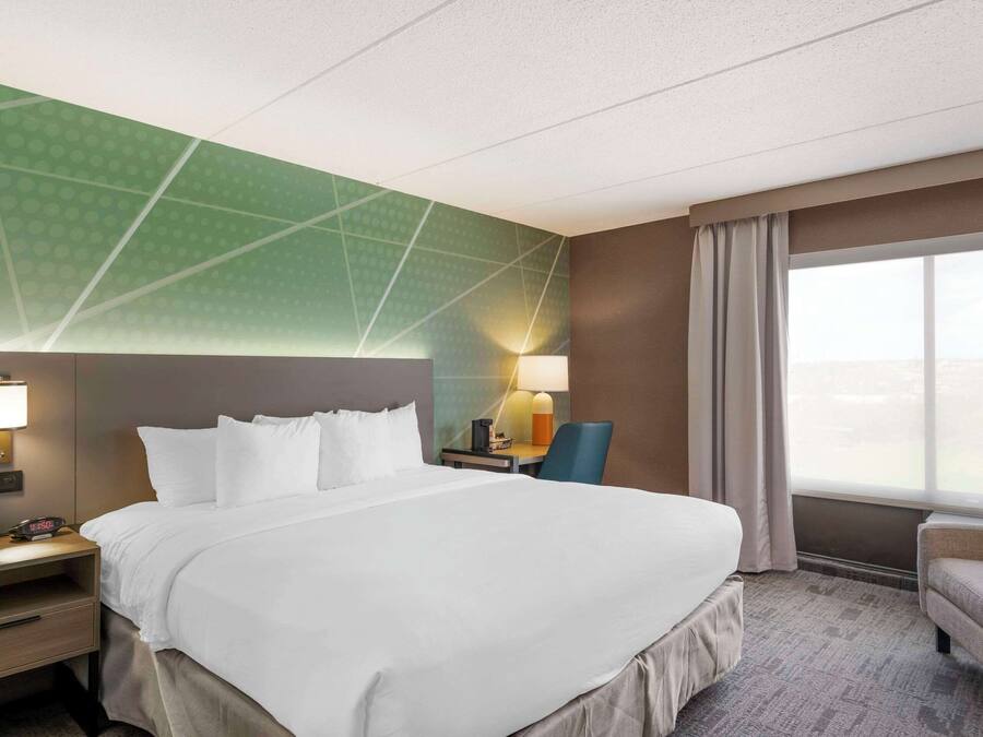 Ein Hotelzimmer mit einem großen Bett, einem Schreibtisch mit Lampe, einem Sessel und Blick auf die Stadt durch das Fenster.