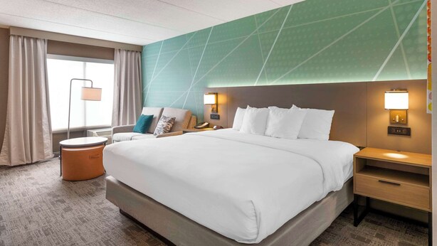 A hotel room with a large bed, a nightstand, a lamp, a chair, and a window with curtains. - Comfort Inn & Suites Watertown - 1000 Islands (Watertown)