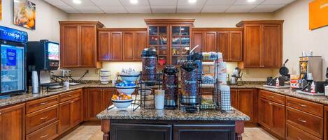 Una cocina de hotel con armarios de madera, encimeras de granito y una estación de café con diferentes cafeteras y recipientes.
