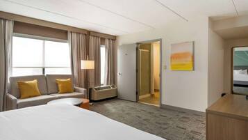 A hotel room with a bed, a sofa with yellow cushions, a desk, a lamp, and a painting on the wall.