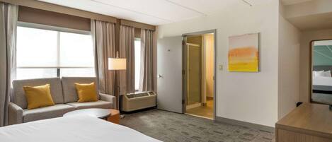 Una habitación de hotel con cama, sofá con cojines amarillos, escritorio, lámpara y un cuadro en la pared.