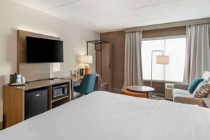 Habitación de hotel con cama, escritorio con televisor de pantalla plana, microondas, refrigerador pequeño, silla, mesa redonda y lámpara.