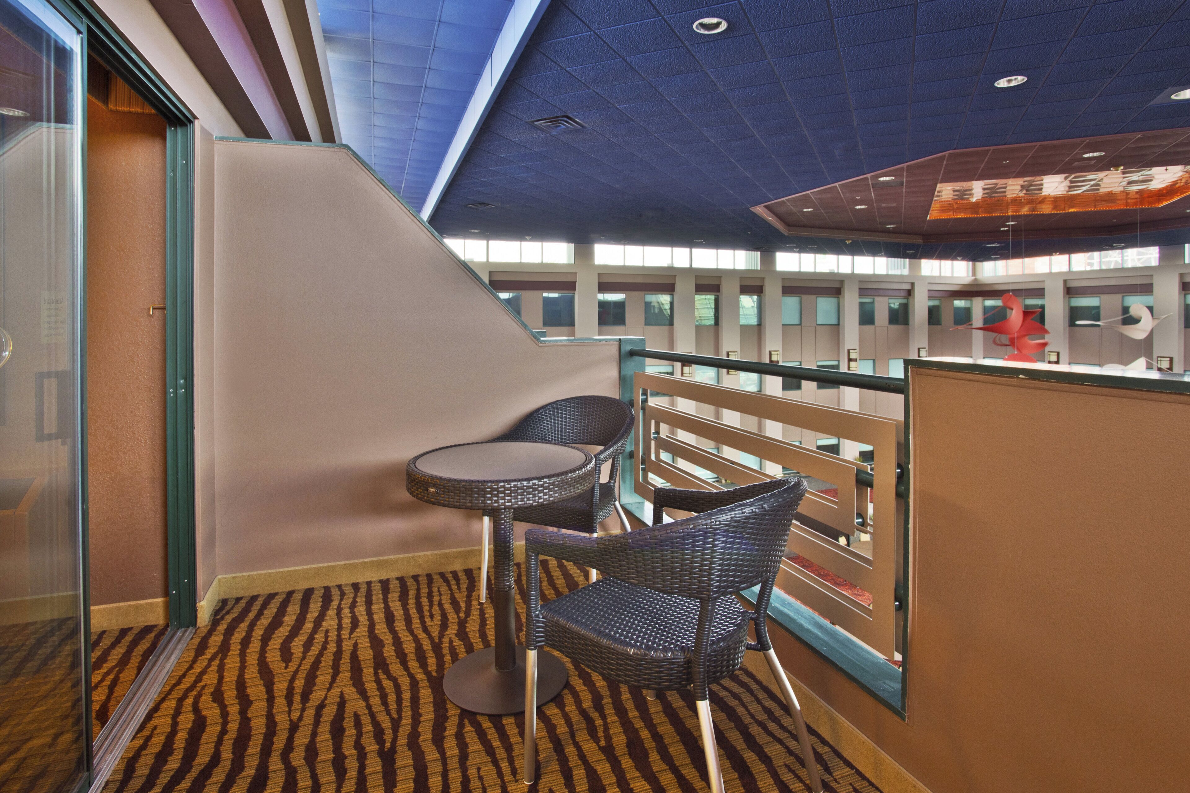 A balcony with two wicker chairs and a small table, a patterned carpet, and a view of the building's interior.