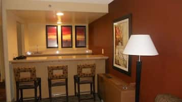 A hotel room with a bar area featuring three chairs, a lamp, and a framed picture on the wall.