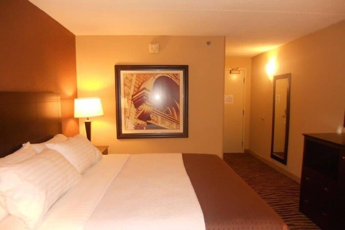 A hotel room with a large bed, a framed picture on the wall, a lamp, and a mirror.