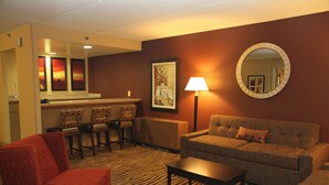 A hotel room with a sofa, armchair, and a small table. There is a mirror on the wall and a lamp. The room has a carpeted floor and a picture of a sunset on the wall.