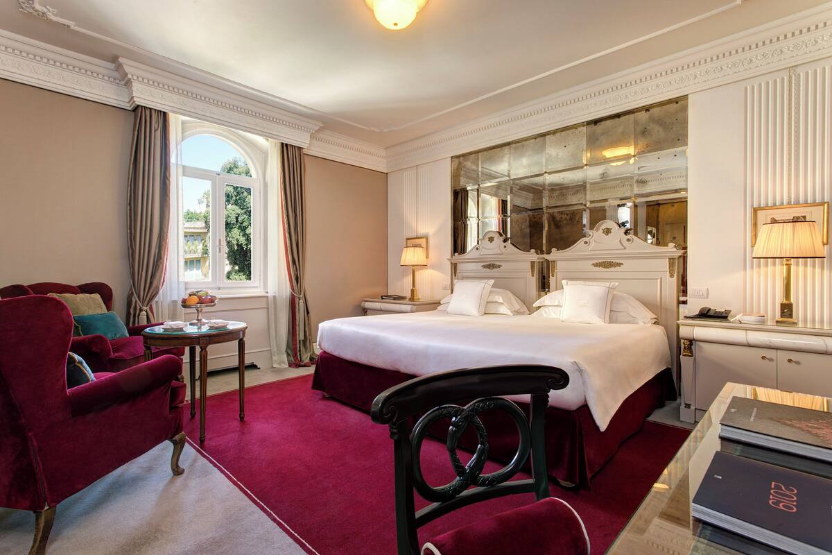 A hotel room with a large bed, a red sofa, a small table, and a mirror.