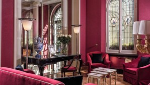 Une pièce comprenant un grand piano, des fauteuils à revêtement rouge, une petite table et une grande fenêtre à vitraux.