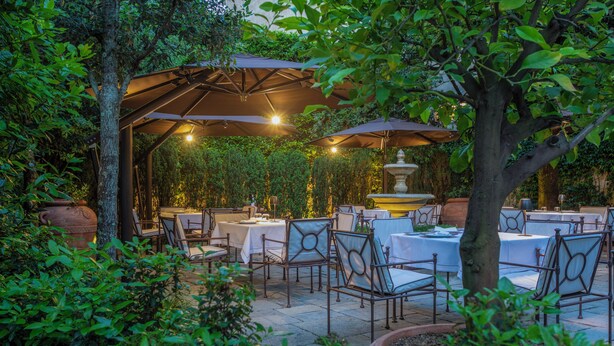 Une terrasse extérieure avec des tables et des chaises, entourée de verdure et agrémentée d’une pergola illuminée.