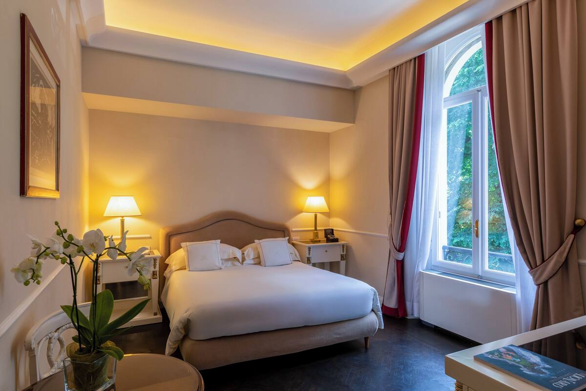 A hotel room with a bed, bedside lamps, a nightstand, a vase with flowers, and a window with curtains.