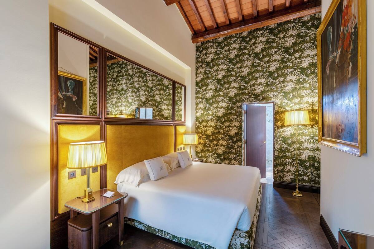 A hotel room with a large bed, two bedside lamps, a nightstand, a mirror, a wardrobe, and a floral wallpaper.