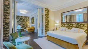 Une chambre d’hôtel avec un grand lit, une table ronde, deux chaises, un miroir et une porte donnant sur une autre pièce.