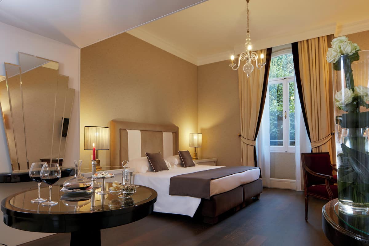 A hotel room with a bed, bedside lamps, a round table with wine glasses, and a vase with flowers.