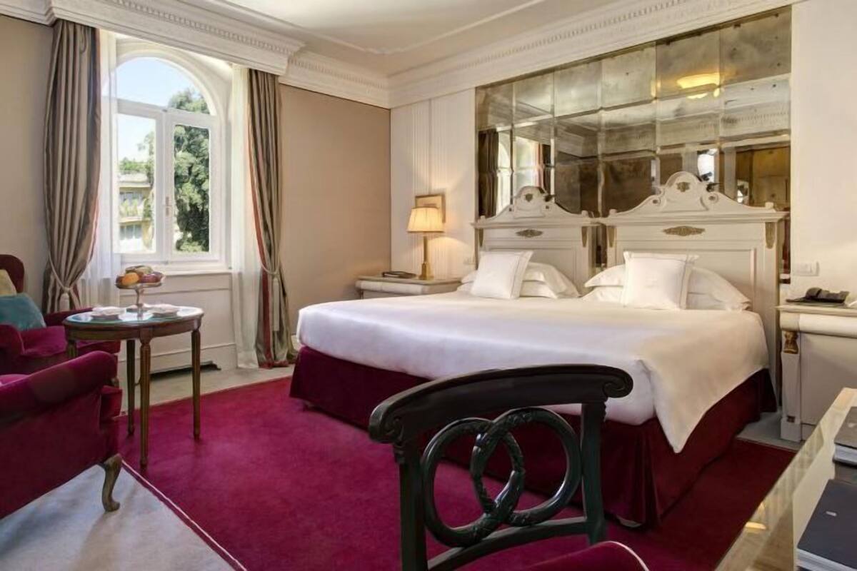 A hotel room with a large bed, a small round table, a chair, a nightstand, and a window with curtains.