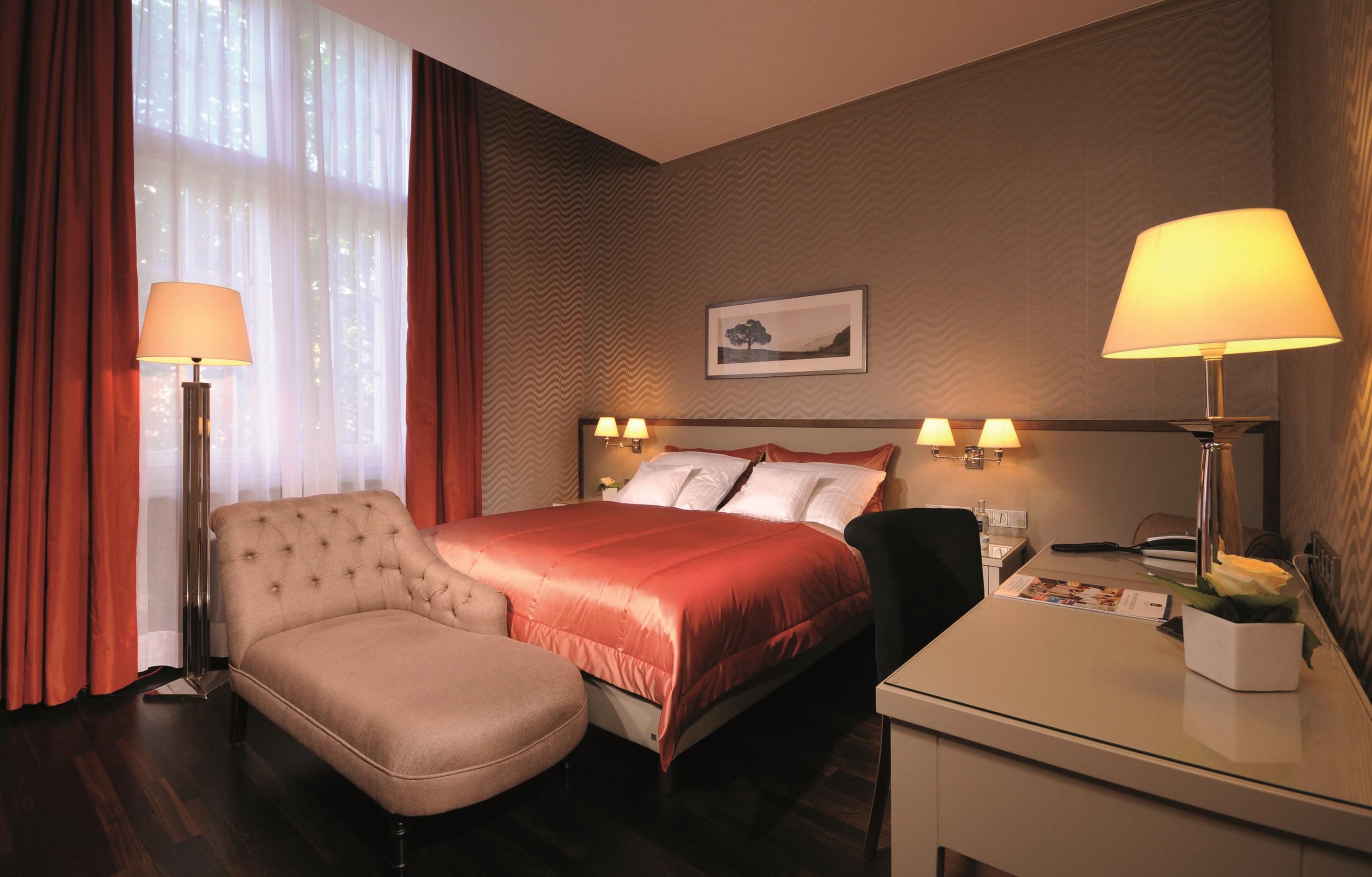 Habitación de hotel con cama, una silla, escritorio, lámparas y un cuadro en la pared.