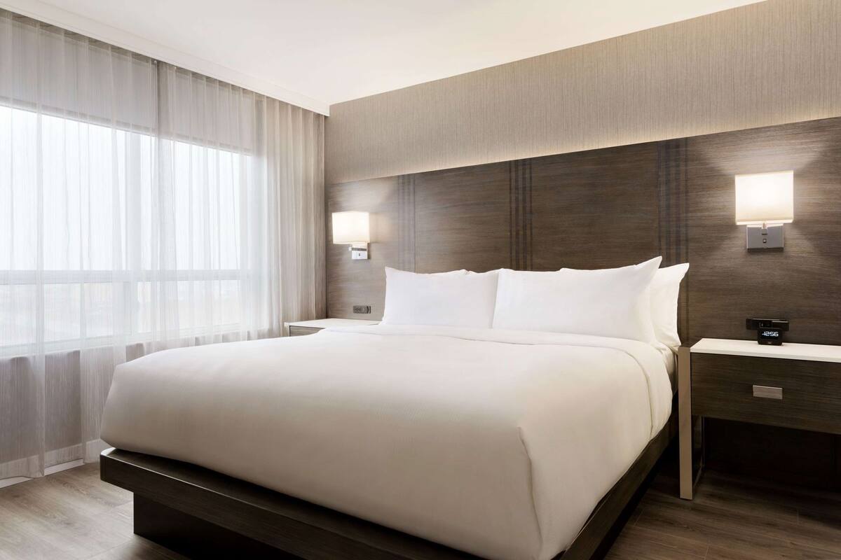 A hotel room with a large bed, two bedside tables, a nightstand, a wall-mounted lamp, and a window with sheer curtains.