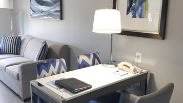 A modern hotel room with a gray sofa, a desk with a lamp, and two framed artworks on the wall.
