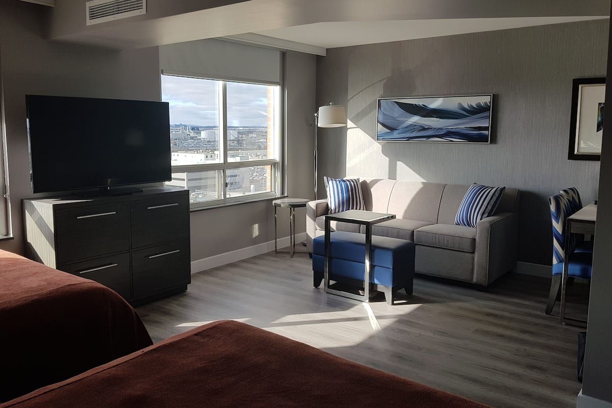 A modern hotel room with a large TV, a sofa, a small table, and a view of the city through large windows.