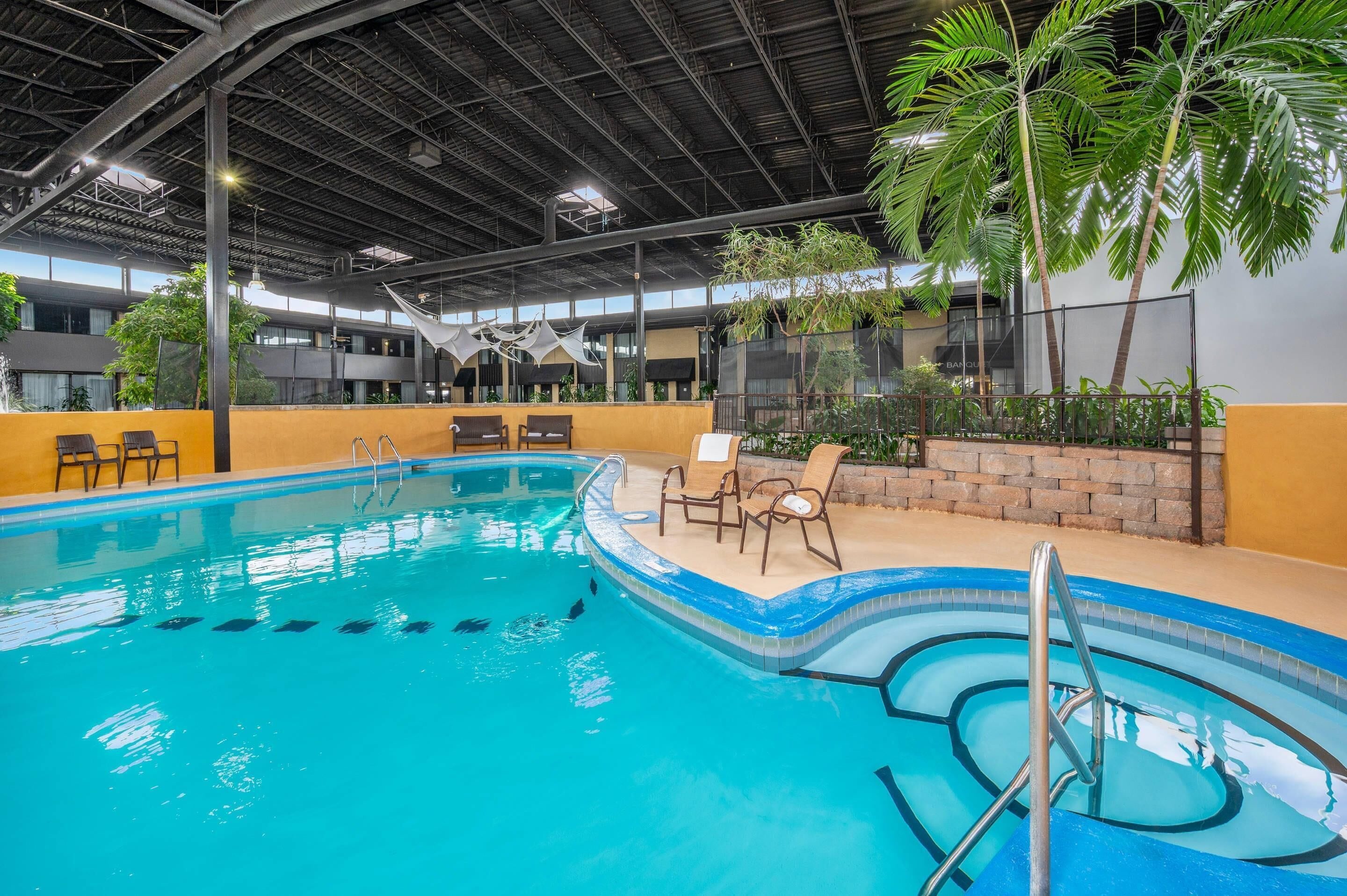 Una piscina interior con agua cristalina, rodeada de sillas de descanso y palmeras.