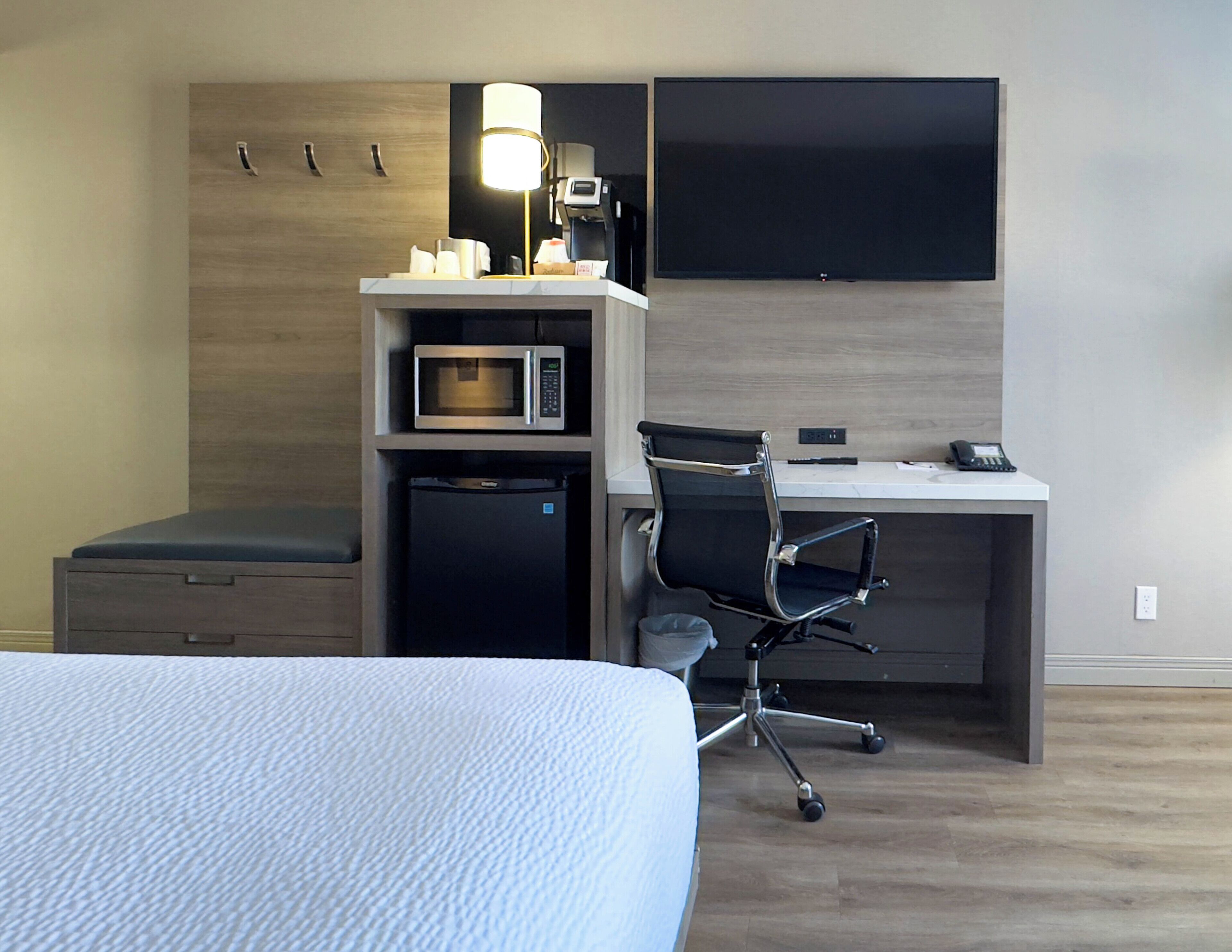 Une chambre d’hôtel équipée d’un lit, d’un bureau avec une chaise, d’un four à micro-ondes, d’un réfrigérateur, d’une lampe et d’un téléviseur à écran plat fixé au mur.