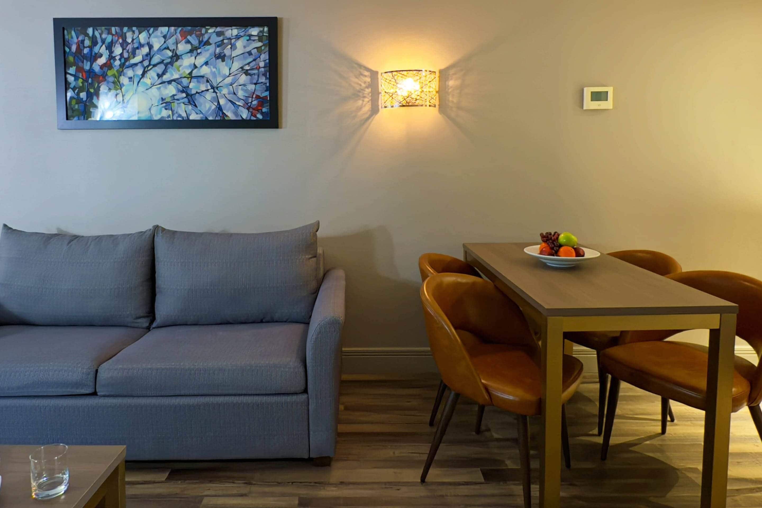A living room with a gray sofa, a wooden dining table with orange chairs, a wall-mounted light fixture, and a framed abstract painting.