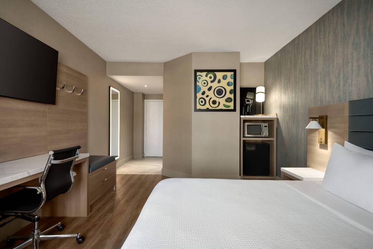 Una habitación de hotel con cama, escritorio con silla, televisor de pantalla plana, microondas, refrigerador y una obra de arte colgada en la pared.