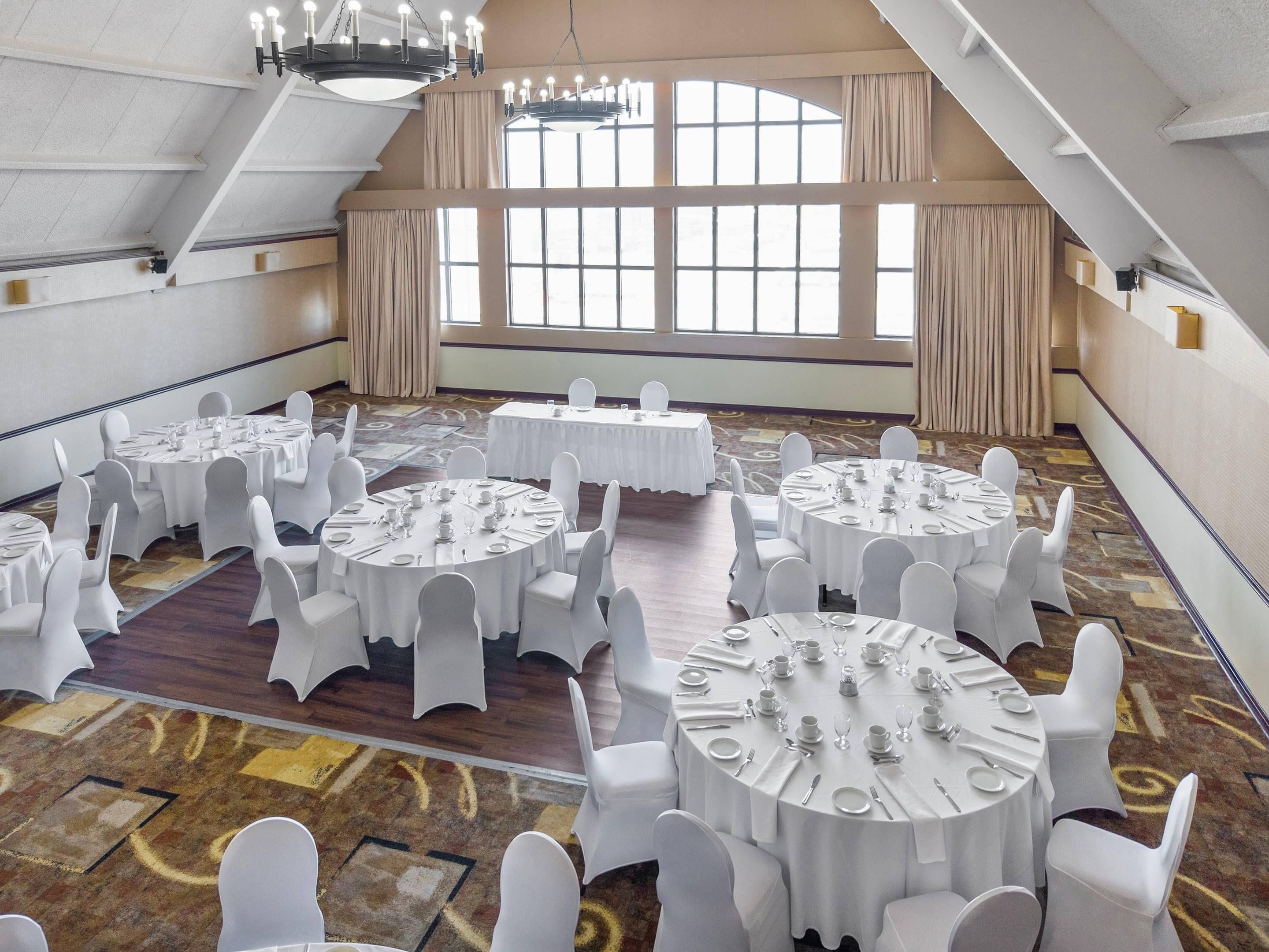 Un amplio y luminoso salón de banquetes con varias mesas redondas dispuestas para cenar, techos altos y grandes ventanales que dejan entrar luz natural.