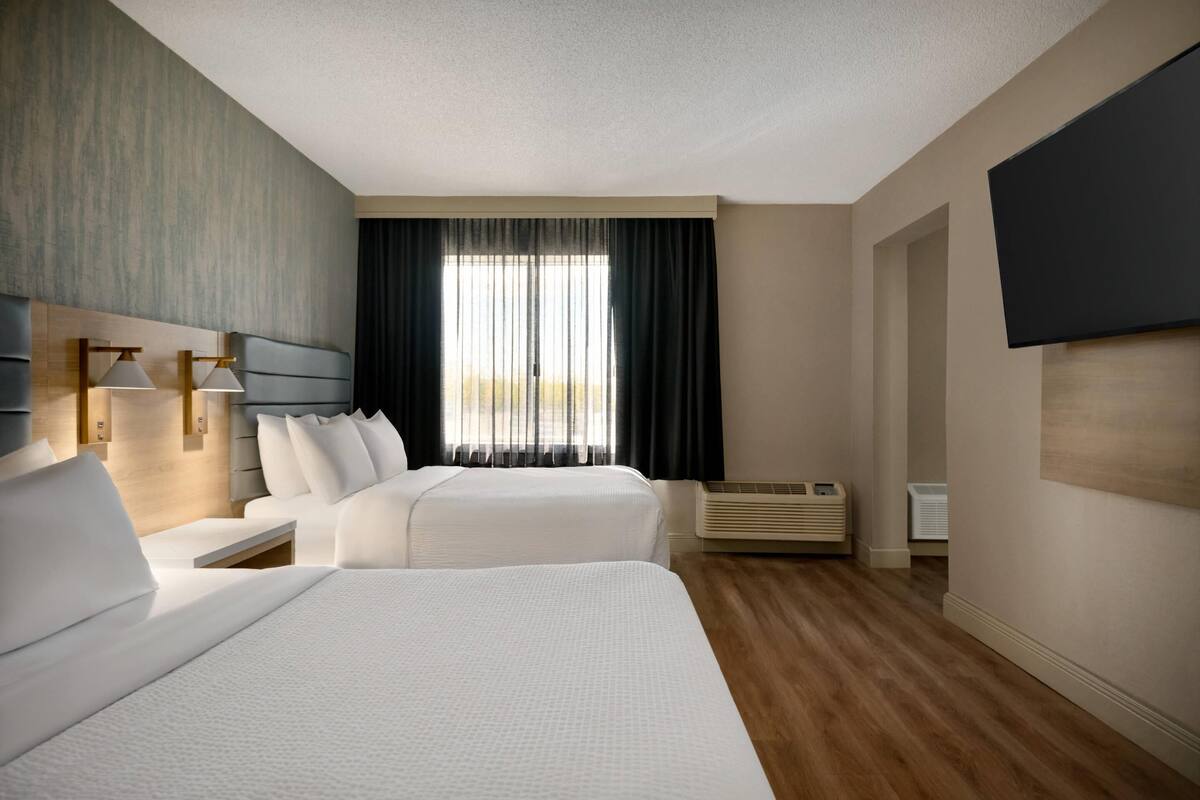Una habitación de hotel moderna con dos camas, un televisor de pantalla plana, un ventanal con cortinas y piso de madera.