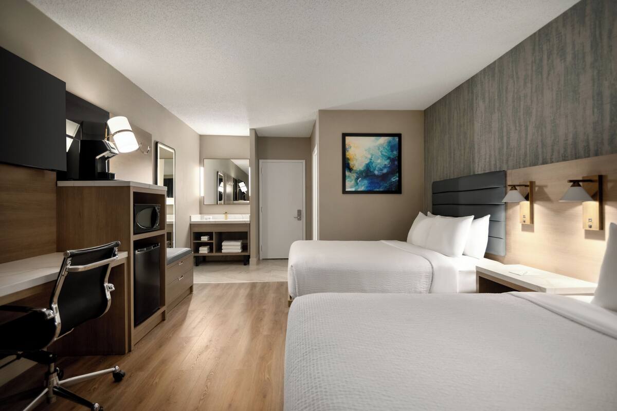 Una habitación de hotel con dos camas, un escritorio con silla, un tocador con espejo y un cuadro en la pared.