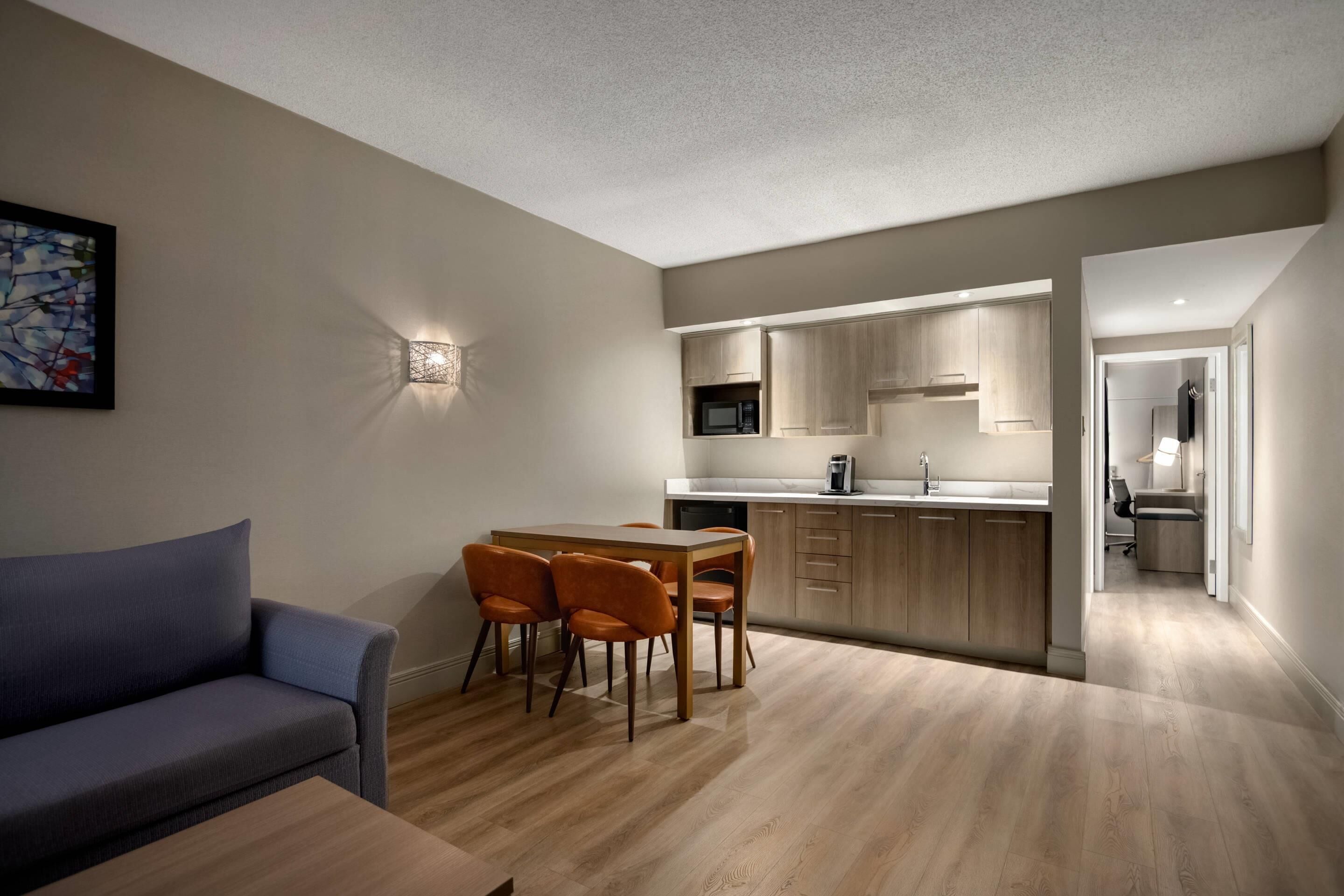 A modern living room with a sofa, a dining table with chairs, a kitchen area with wooden cabinets and a microwave, and a hallway leading to another room.