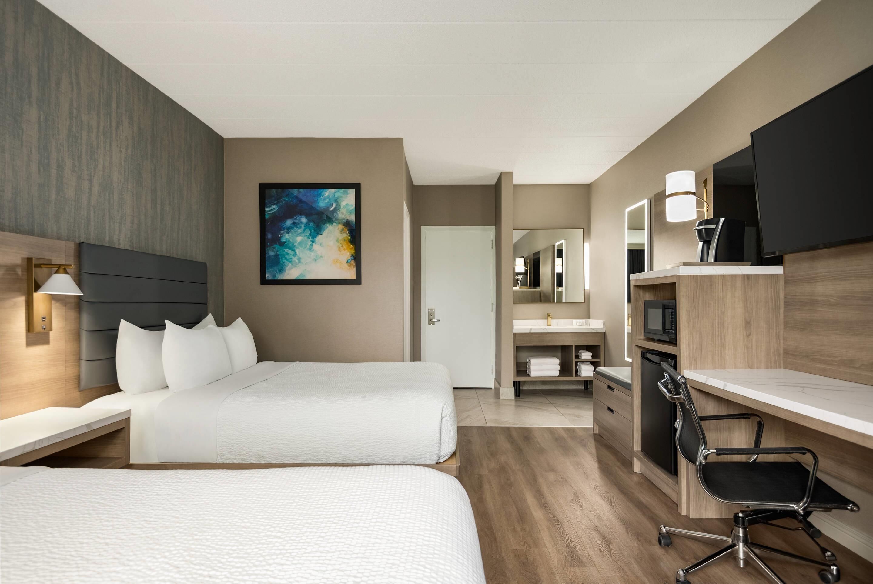 Habitación de hotel con dos camas, un escritorio, una silla, televisor, microondas y baño con espejo y lavamanos.
