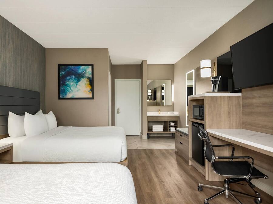 Habitación de hotel con dos camas, un escritorio, una silla, televisor, microondas y baño con espejo y lavamanos.