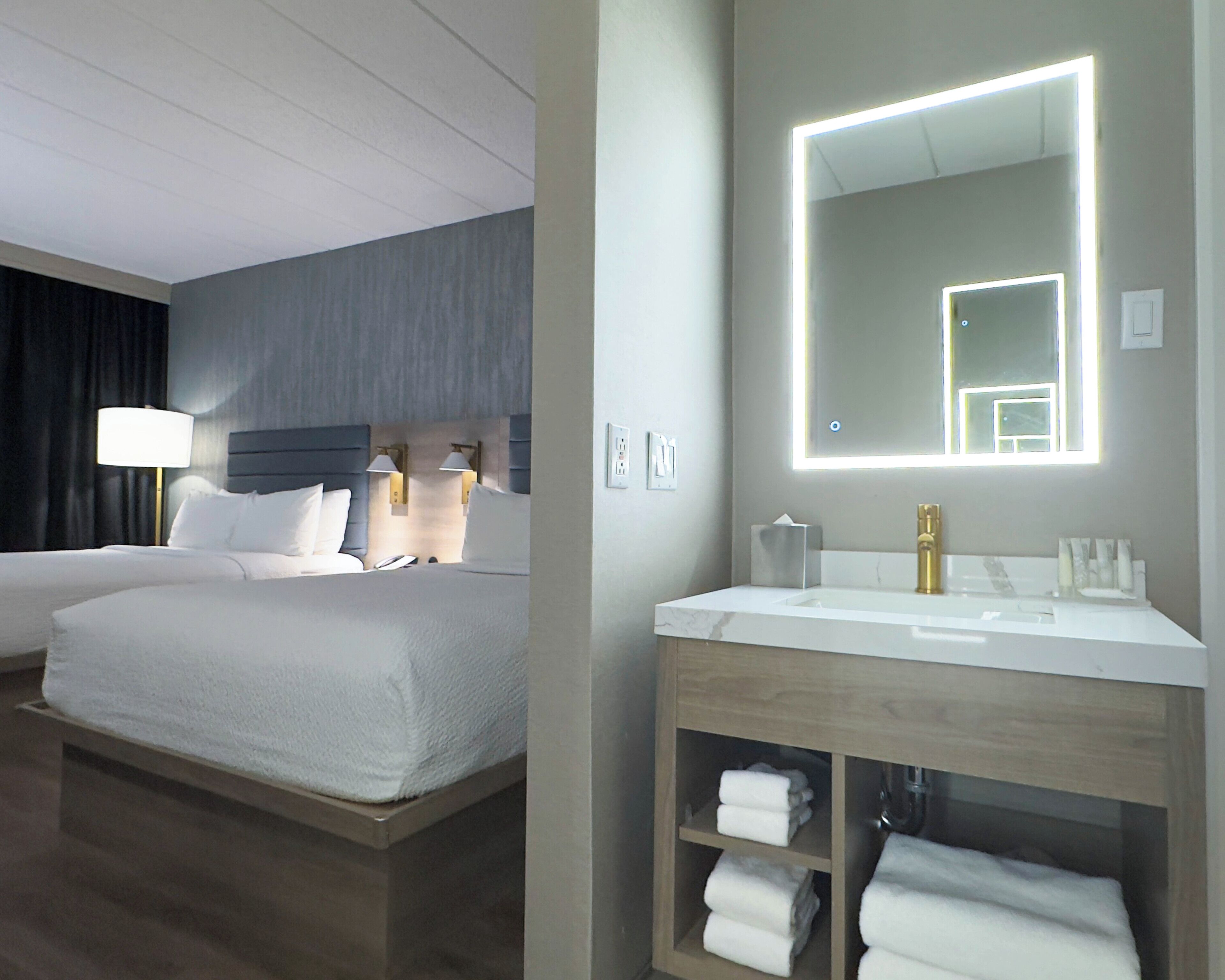 A hotel room with a large bed, bedside lamps, a vanity with a mirror, and a shelf with folded towels.
