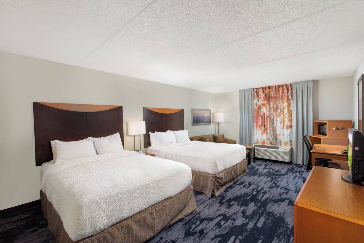 A hotel room with two beds, a desk, a TV, and a window with curtains.