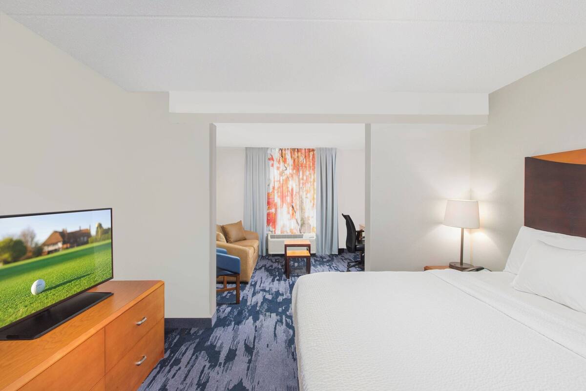 A hotel room with a bed, a TV on a wooden dresser, a desk with a chair, and a lamp.